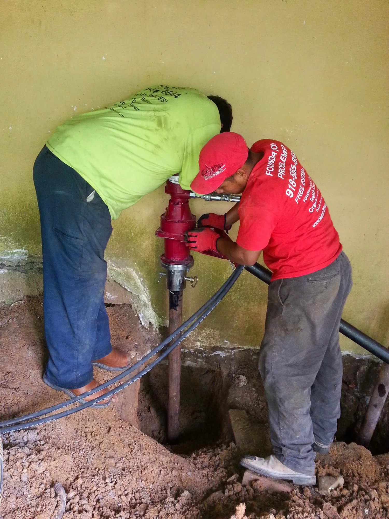 Two technicians installing red fire hydrant at foundation access point with excavated pit
