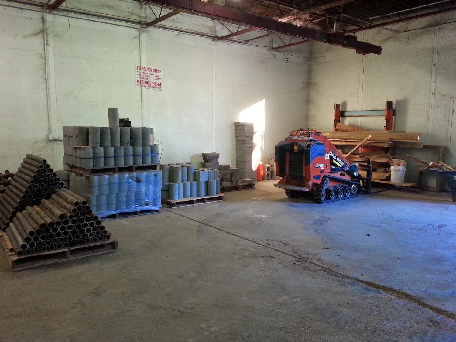 Industrial warehouse with stacked concrete pipes, blue drums, and foundation repair equipment staged for project work