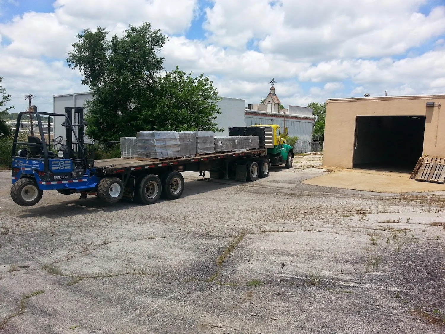 Blue forklift loading wrapped pallets onto flatbed truck at industrial yard