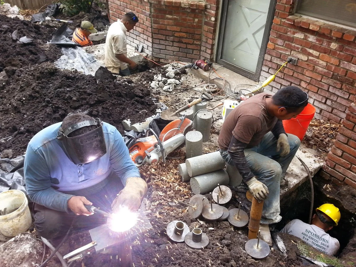 Foundation repair crew installing steel piers and concrete footings against brick foundation