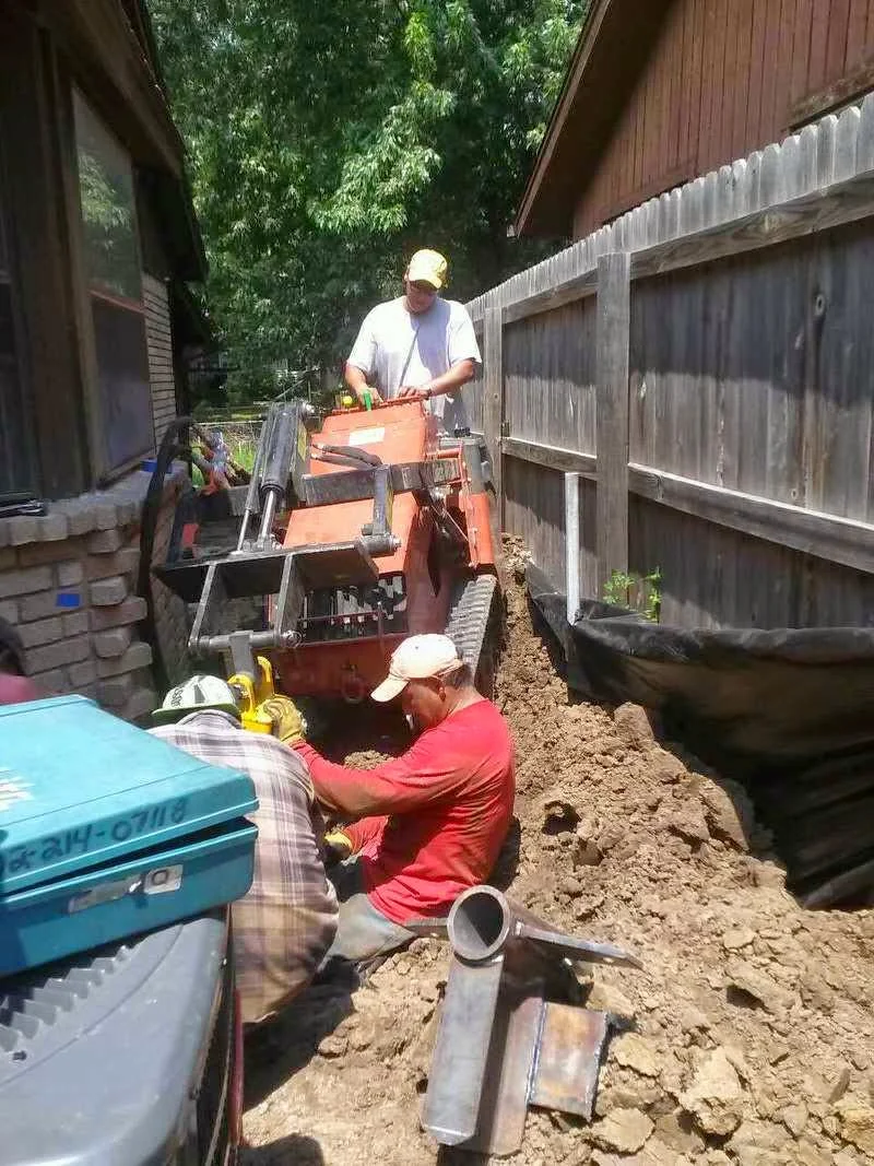Foundation drainage installation with excavation equipment and crew working in tight residential space