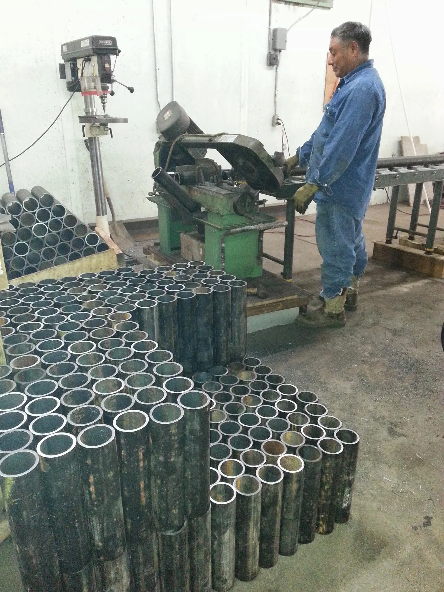 Steel foundation piers with welded caps staged in workshop, worker operating drilling equipment